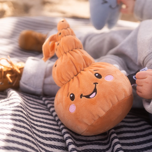 Ein Kind sitzt auf einer gestreiften Decke und hält Mia, die weiche Plüschmuschel. Mia ist warm braun, hat mehrere sanft geschichtete Muschelsegmente und ein freundliches Gesicht mit rosa Wangen. Die Szene ist in weiches Sonnenlicht getaucht.